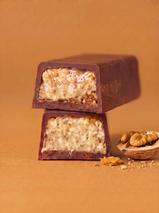 Two stacked cross-section pieces of the Après l'Étoile Walnut + Cinnamon Baklava Milk Chocolate Bar, revealing a flaky, nut-studded baklava filling encased in milk chocolate, with a cracked walnut shell and pastry flakes beside them on a warm amber background.