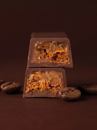 Two stacked cross-section pieces of the Après l'Étoile Coffee Caramel + Pecan Dark Chocolate Bar, revealing a rich coffee caramel and pecan filling encased in dark chocolate, surrounded by whole coffee beans on a deep brown background.
