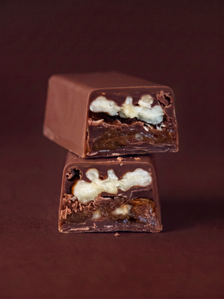 Two halves of a chocolate bar stacked vertically, showing a rich dark chocolate filling with white chocolate drizzle and chopped nuts, set against a dark brown background.