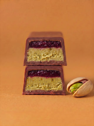 wo stacked cross-section pieces of the Après l'Étoile Raspberry Tea + Pistachio Milk Chocolate Bar, revealing layers of pistachio cream and raspberry jam encased in milk chocolate, with a cracked pistachio shell beside them on a warm amber background