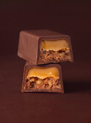 Two stacked cross-section pieces of the Après l'Étoile Sticky Toffee Milk Chocolate Bar, revealing a golden toffee caramel layer over a dense sticky toffee filling, all encased in smooth milk chocolate, set against a deep brown background.